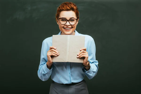 Attractive, happy teacher in eyeglasses holding open book while standing near chalkboard — Stock Photo