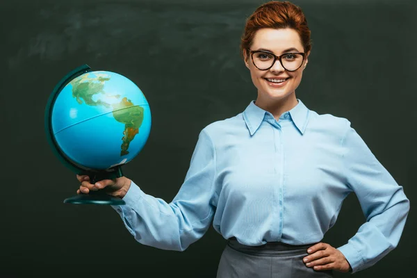 Happy young teacher standing near chalkboard with hand on hip and holding globe — Stock Photo
