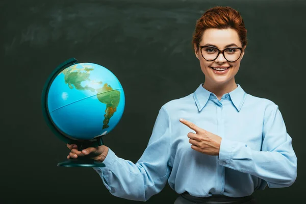 Smiling teacher pointing with finger at globe while standing near chalkboard — Stock Photo