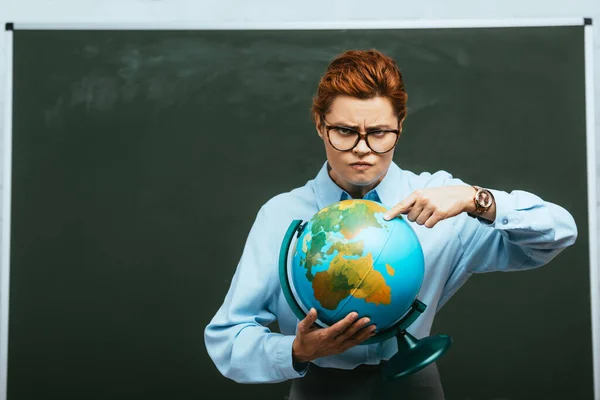 Strict teacher pointing with finger at globe while standing near chalkboard — Stock Photo