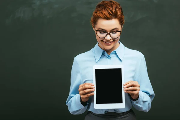 Smiling teacher holding digital tablet with blank screen while standing near chalkboard — Stock Photo