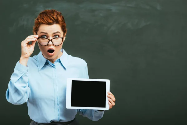 Шокований вчитель торкається окулярів, тримаючи цифровий планшет з порожнім екраном біля крейдяної дошки — Stock Photo