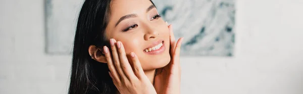 Panoramaaufnahme einer lächelnden asiatischen Frau, die ihr Gesicht zu Hause berührt — Stockfoto