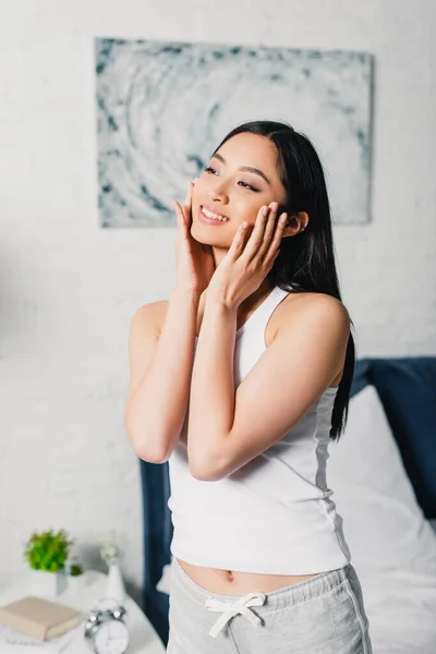 Schöne asiatische Frau lächelt, während sie ihr Gesicht zu Hause berührt — Stockfoto