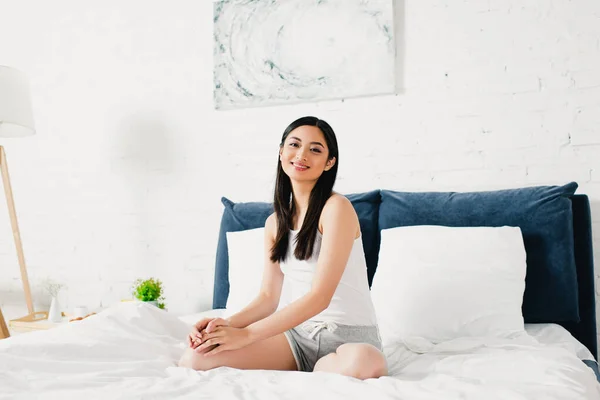 Красива азіатська жінка посміхається на камеру, сидячи на ліжку — стокове фото