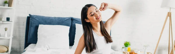 Panoramic shot of smiling asian woman stretching on bed during morning — Stock Photo