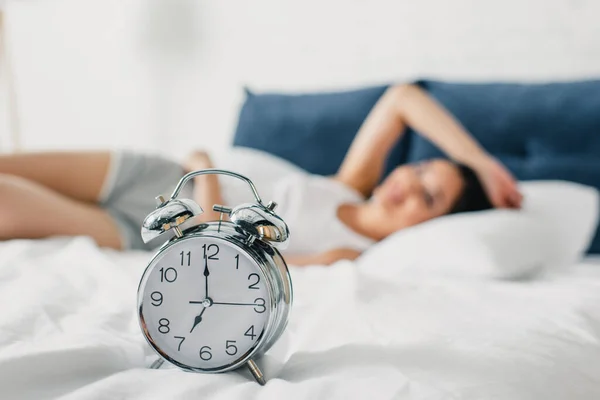 Selective focus of alarm clock on bedding and young woman lying on bed at morning — Stock Photo