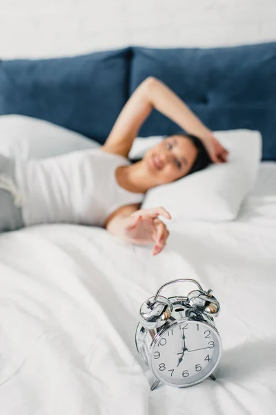 Focus selettivo della sveglia sulla biancheria da letto e sulla donna che tira la mano sul letto al mattino — Foto stock