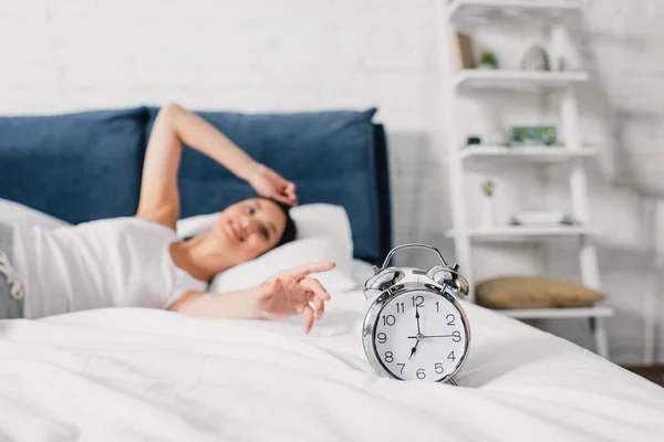 Вибірковий фокус усміхненої азіатки тягне руку на будильник на ліжку вранці — стокове фото