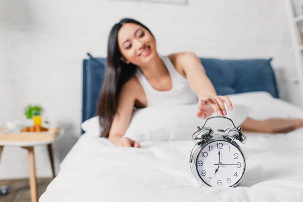 Selective focus of alarm clock on bedding and cheerful asian girl pulling hand while lying on bed — Stock Photo