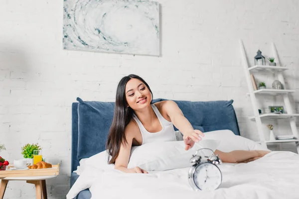 Foco seletivo de bela mulher asiática sorrindo e puxando a mão para despertador na cama de manhã — Fotografia de Stock