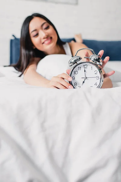 Selective focus of smiling asian woman holding alarm clock on bed at morning — Stock Photo