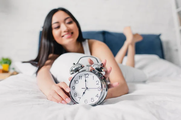 Enfoque selectivo de positivo asiático chica sosteniendo alarma reloj mientras está acostado en la cama por la mañana - foto de stock