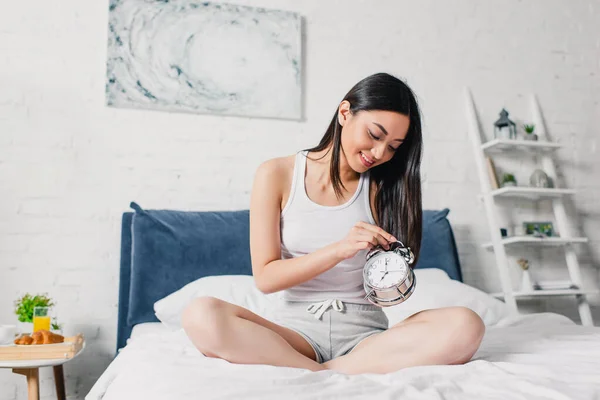 Linda menina asiática segurando despertador e sorrindo na cama de manhã — Fotografia de Stock