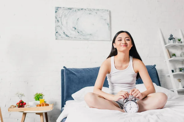 Feliz asiático mujer sonriendo en cámara cerca alarma reloj en cama en la mañana - foto de stock