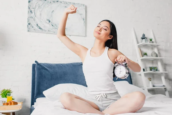 Beautiful asian woman stretching with closed eyes while holding alarm clock on bed at morning — Stock Photo