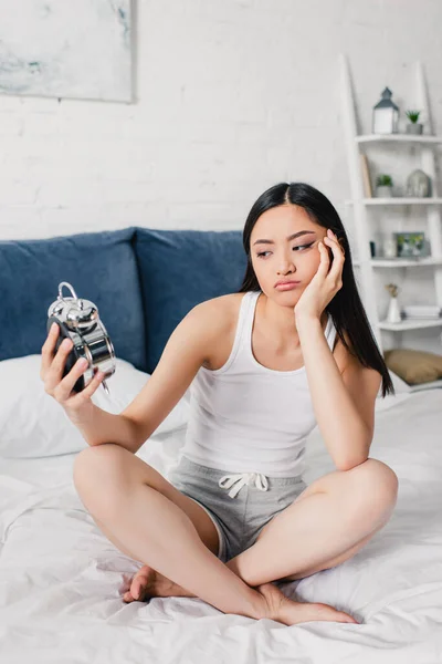 Skeptisch asiatisch mädchen looking at wecker while sitting on bed at morning — Stockfoto