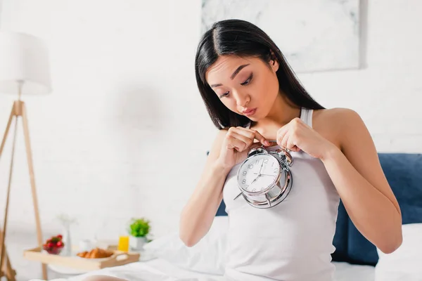 Mujer asiática sorprendida mirando el reloj despertador en el dormitorio por la mañana - foto de stock