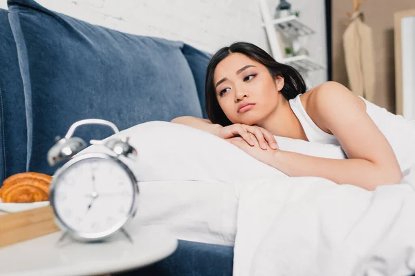 Selective focus of sad asian girl lying on bed near alarm clock and breakfast on bedside table — Stock Photo