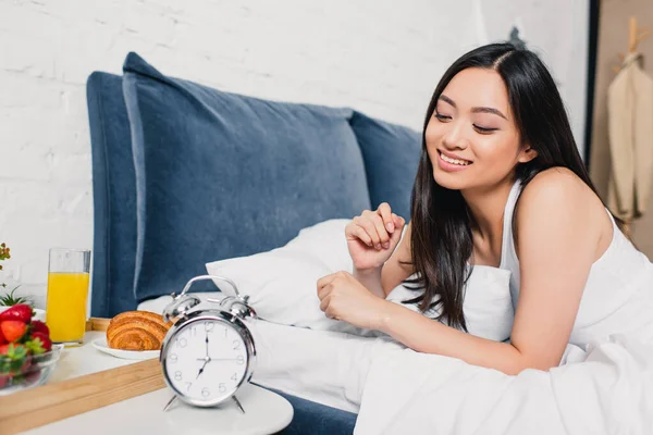 Selective focus of smiling asian girl lying on bed near breakfast and alarm clock on bedside table — Stock Photo