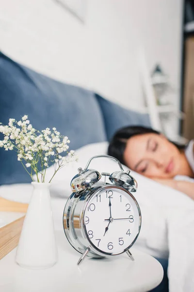 Вибірковий фокус будильника і квіти у вазі на тумбочці і азіатська жінка спить на ліжку вранці — стокове фото