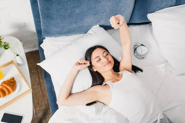 Top view of asian girl stretching near alarm clock, smartphone and breakfast on bedside tale — Stock Photo