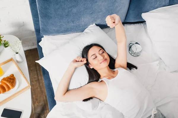 Vista dall'alto della ragazza asiatica che si estende vicino alla sveglia, smartphone e colazione sul comodino — Foto stock