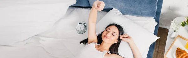 Panoramic shot of asian girl stretching near alarm clock and breakfast on bedside table — Stock Photo