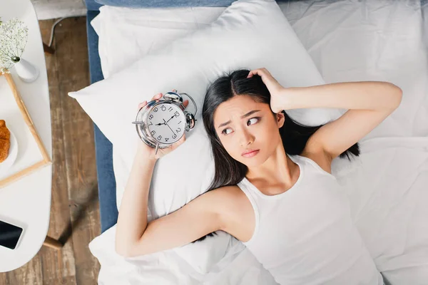 Vista superior de la triste mujer asiática sosteniendo reloj despertador en la cama por la mañana - foto de stock