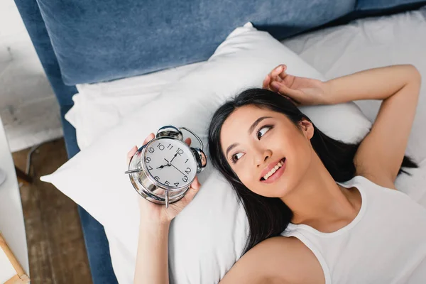 Top vista do sorriso asiático mulher olhando para despertador enquanto deitado na cama de manhã — Fotografia de Stock