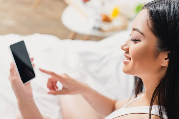 Foco seletivo de sorrir menina asiática apontando com o dedo para o smartphone na cama — Fotografia de Stock