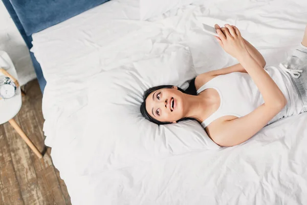 Vista dall'alto della donna asiatica sorridente che tiene lo smartphone e distoglie lo sguardo sul letto al mattino — Foto stock