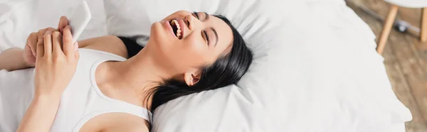 Panorâmica tiro de mulher asiática rindo enquanto segurando smartphone na cama — Fotografia de Stock