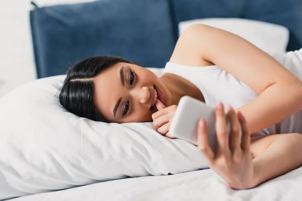 Enfoque selectivo de sonreír mujer asiática con el dedo cerca de los labios utilizando el teléfono inteligente en la cama - foto de stock