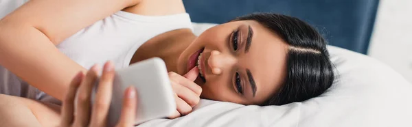 Panoramic shot of smiling asian woman with finger near lips using smartphone on bed — Stock Photo