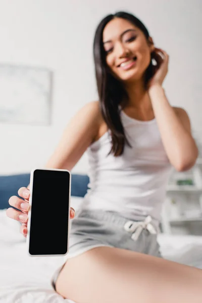 Selective focus of smiling asian woman showing smartphone with blank screen on bed — Stock Photo