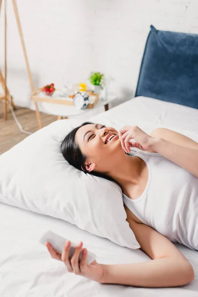 Selective focus of positive asian girl holding smartphone on bed at morning — Stock Photo