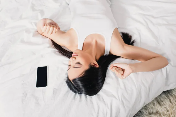 Vista superior de la mujer asiática estiramiento cerca de teléfono inteligente en la cama - foto de stock