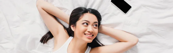 Горизонтальне зображення усміхненої азіатки, дивлячись на смартфон з порожнім екраном на ліжку — стокове фото