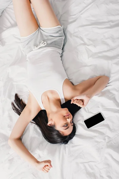 Top view of cheerful asian girl stretching and looking at smartphone on bed — Stock Photo