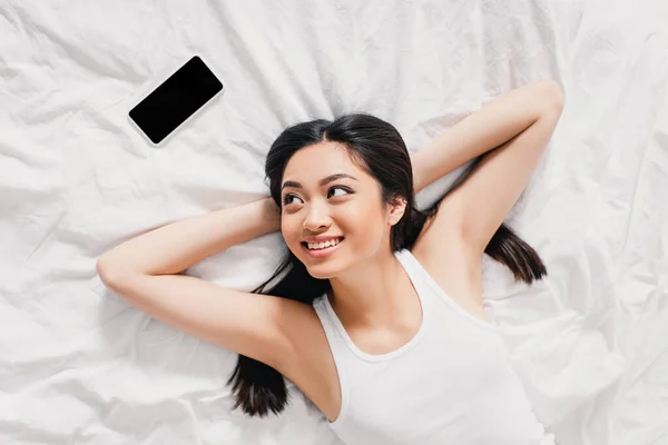 Vista dall'alto di allegra ragazza asiatica in camicia senza maniche guardando smartphone con schermo bianco sul letto — Foto stock