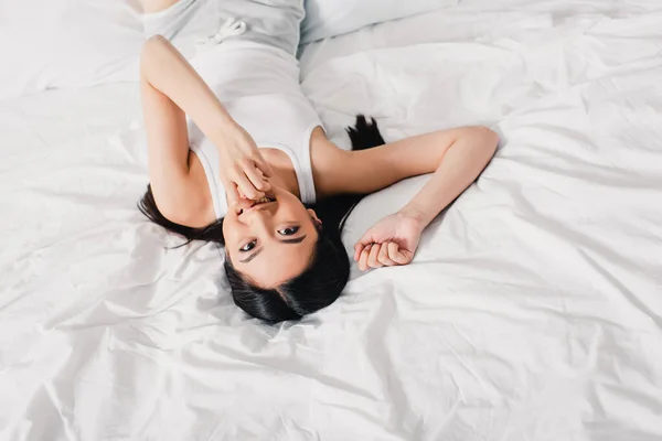 Vue grand angle de fille asiatique avec la main près de la bouche souriant à la caméra sur le lit — Photo de stock