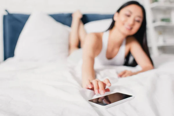 Selective focus of asian woman pulling hand to smartphone on bed at morning — Stock Photo