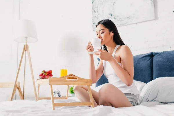 Азиатка пьет кофе у подноса для завтрака на кровати — стоковое фото