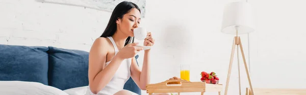 Panoramic shot of asian woman drinking coffee near breakfast tray on bed — Stock Photo
