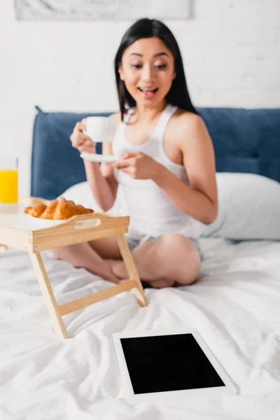 Foco seletivo de mulher asiática positiva segurando xícara de café e olhando para tablet digital na cama — Fotografia de Stock