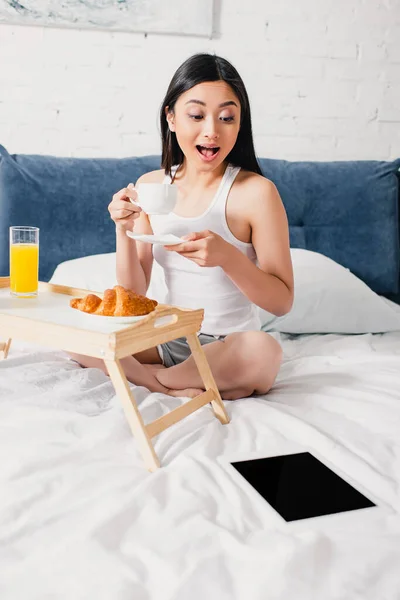 Захоплена азіатська жінка дивиться на цифровий планшет під час пиття кави на ліжку — стокове фото