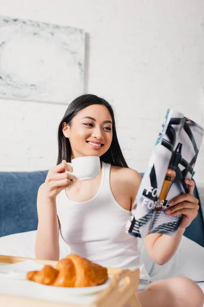 Selektiver Fokus einer lächelnden asiatischen Frau, die eine Tasse Kaffee in der Hand hält und Magazin auf dem Bett liest — Stockfoto