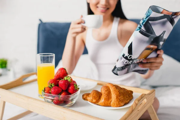 Concentration sélective du croissant, des fraises et du jus d'orange sur le plateau de petit-déjeuner et fille souriante tenant une tasse de café et un magazine au lit — Photo de stock