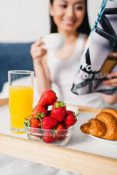 Selective focus of breakfast on tray and woman holding cup of coffee and magazine on bed — Stock Photo